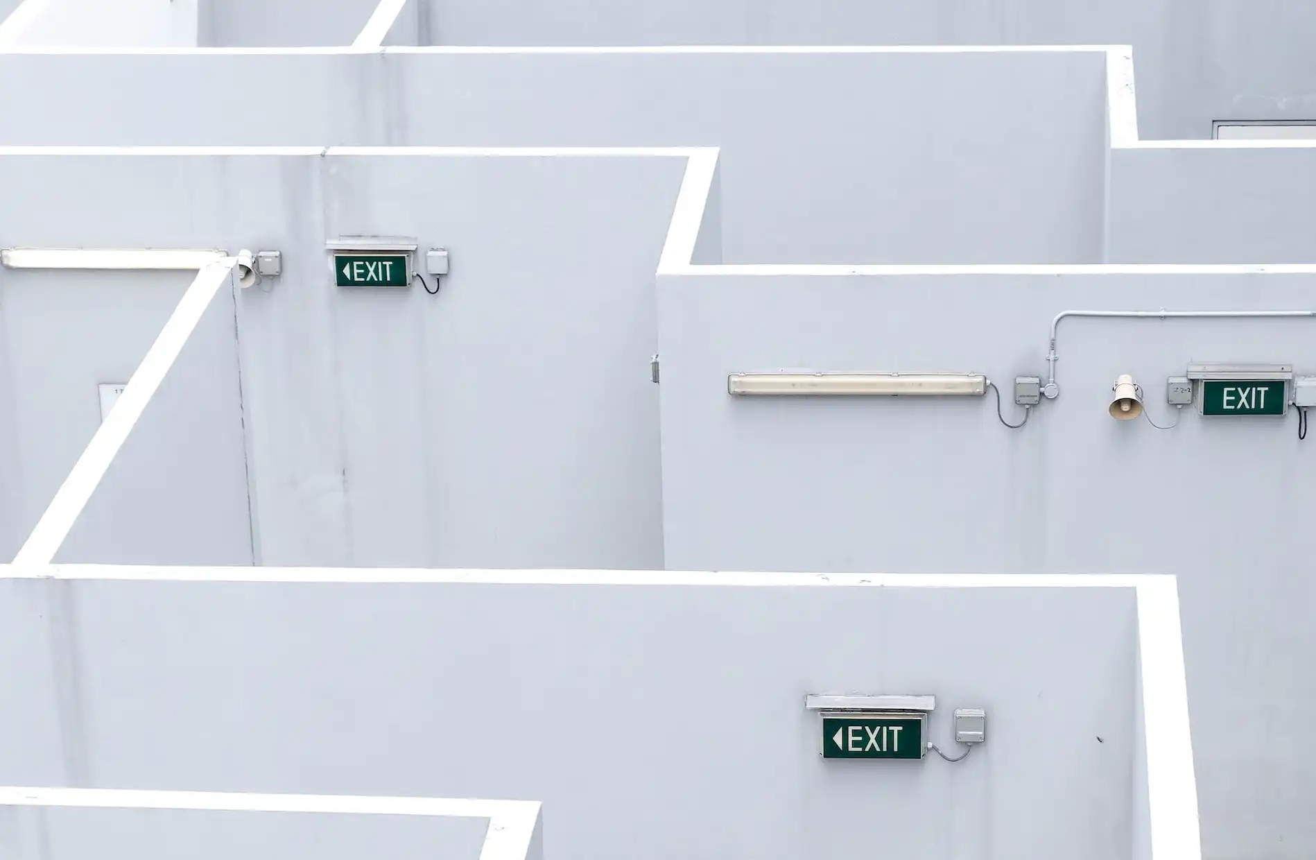 Weiße Wände eines Labyrinths mit mehreren "EXIT"-Schildern, Neonbeleuchtung und Lautsprecher, ohne sichtbare Menschen.