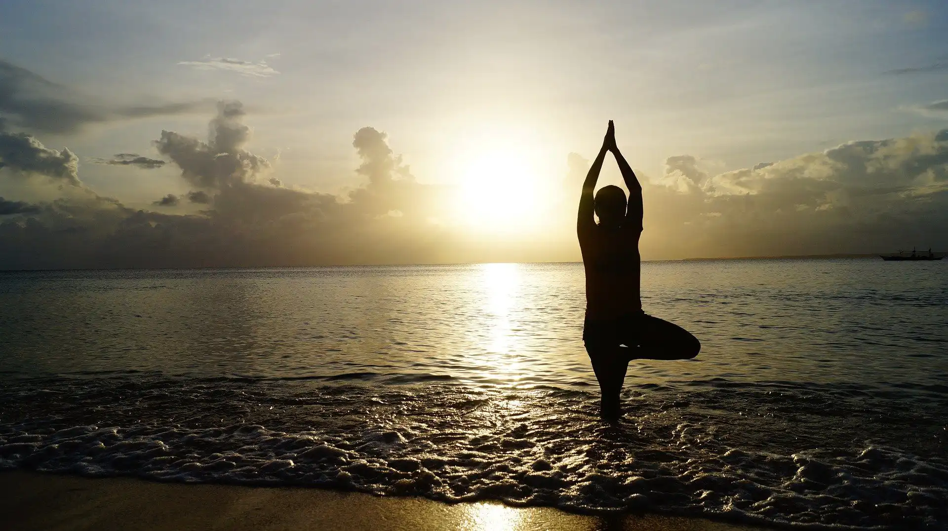 Eine Person steht im Sonnenuntergang am Meer in der Yoga-Baumhaltung, umgeben von sanften Wellen und dramatischen Wolken.