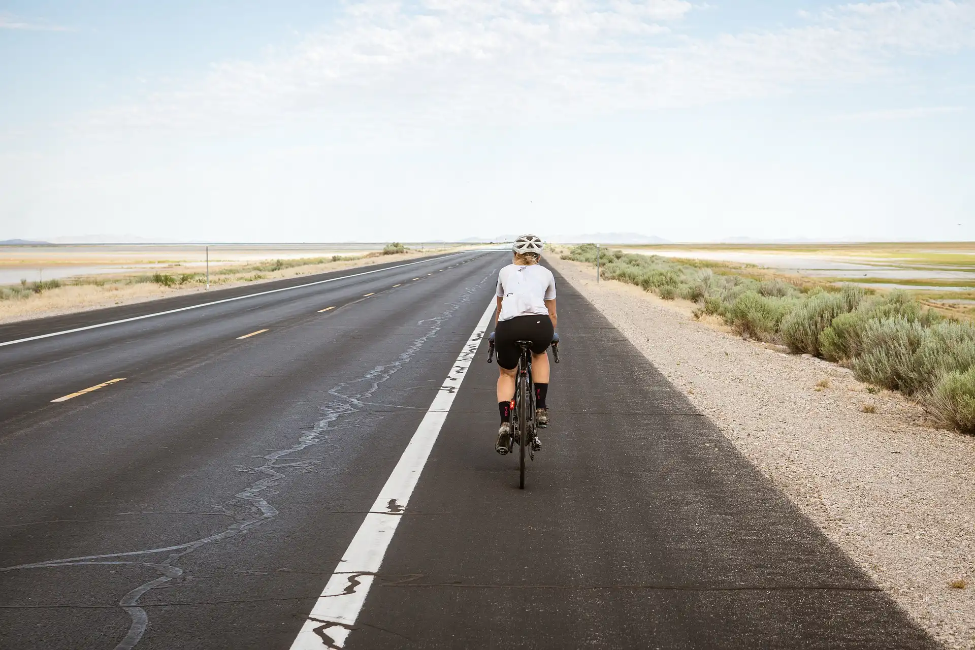 Eine Person fährt mit dem Fahrrad auf einer leeren, geraden Straße durch eine weite, ländliche Landschaft unter einem klaren Himmel.