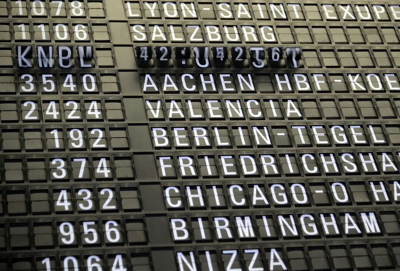 Eine Anzeigetafel mit Zielen wie Lyon, Salzburg, Aachen und Valencia in einer Bahnhof- oder Flughafen-Umgebung zeigt Abfahrts- oder Ankunftsinformationen in leuchtenden Buchstaben.