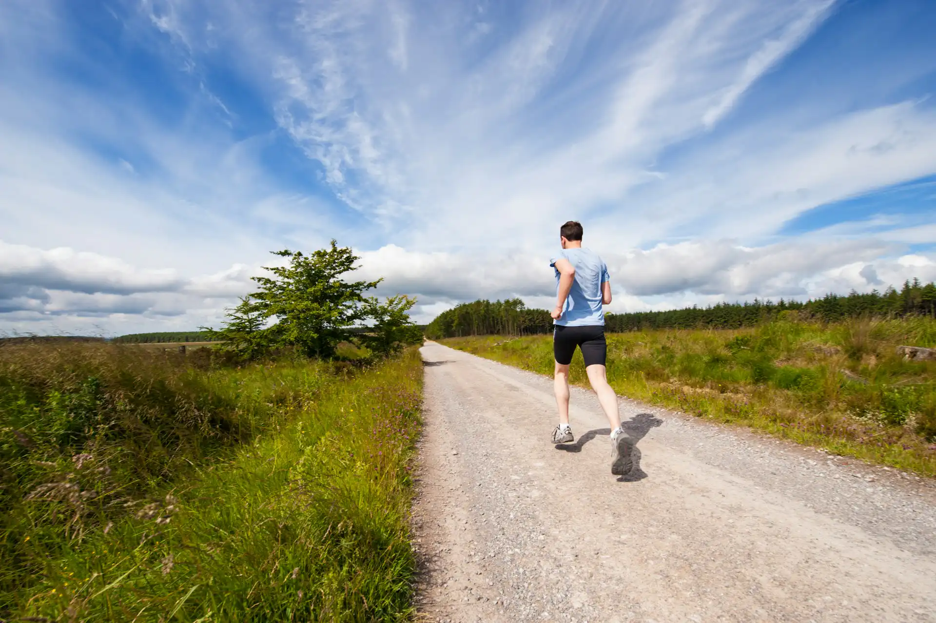 Ein Mann joggt auf einem Kiesweg durch eine ruhige, grüne Landschaft unter einem wolkigen, blauen Himmel.