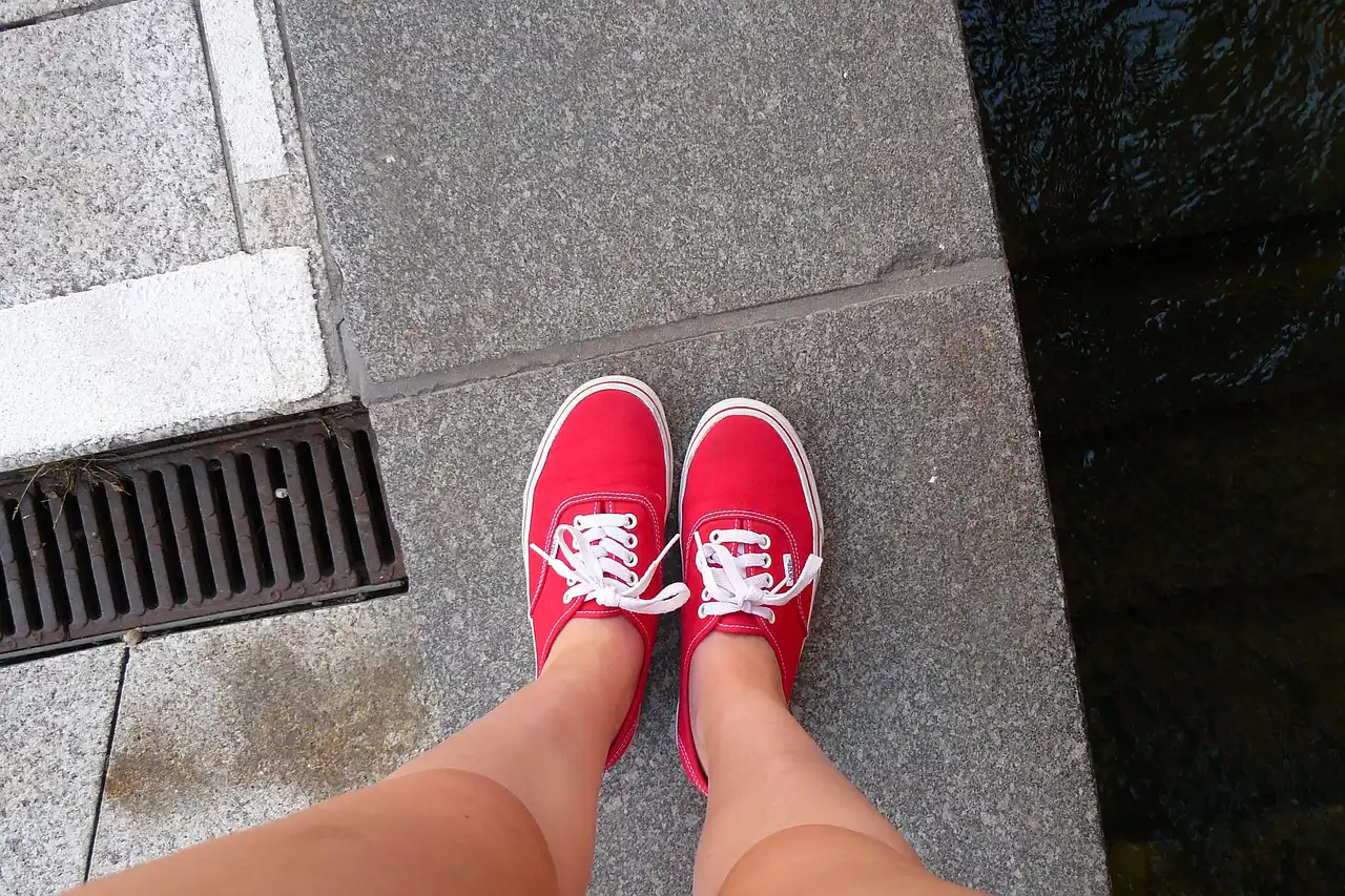 Rote Turnschuhe stehen auf einem grauen Gehweg neben einem Abflussgitter, mit Wasser, das rechts im Bild fließt.