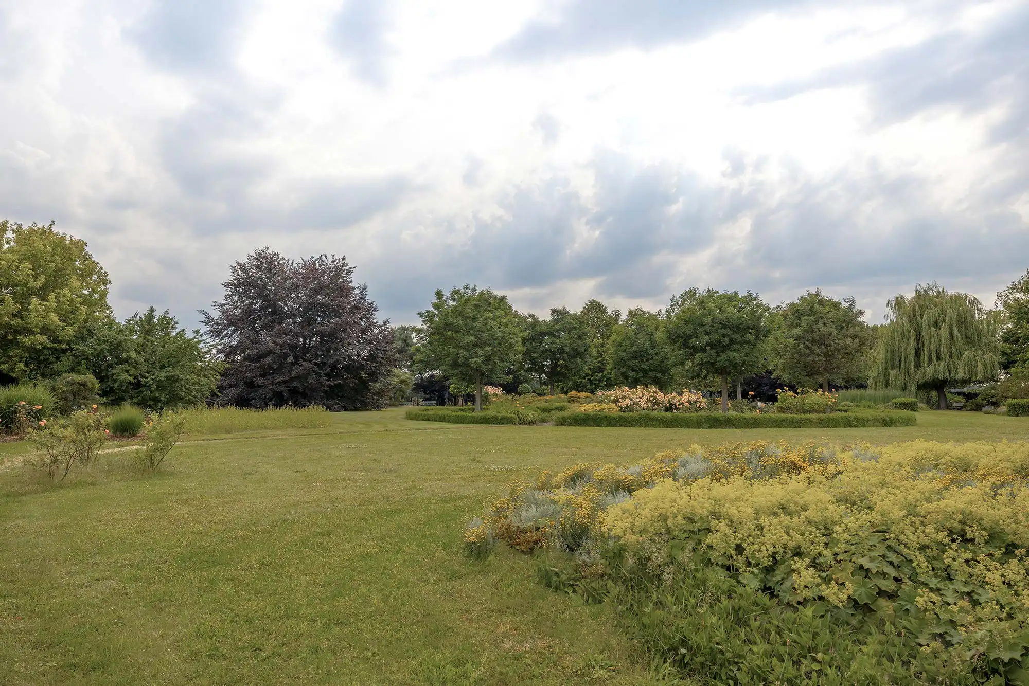 Parkanlage der Privatklinik Merbeck Ein weitläufiger Garten mit gepflegtem Rasen, blühenden Büschen und großen Bäumen unter einem bewölkten Himmel.