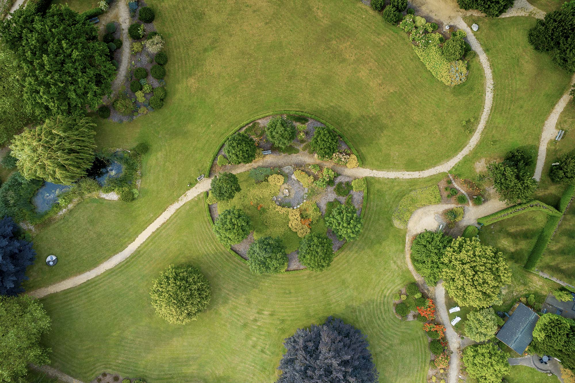 Eine kreisförmige Gartenanlage mit Wegen, Büschen und Bäumen, umgeben von gepflegtem Rasen, bietet eine ruhige Atmosphäre für Entspannung und Erholung in einer privaten Klinik.