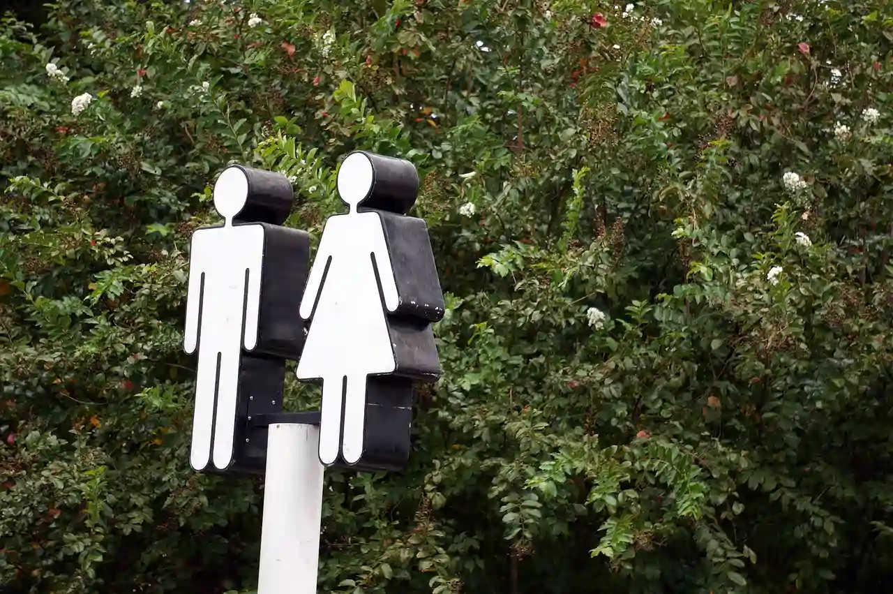 Zwei stilisierte menschliche Figuren aus Metall stehen auf einem Pfosten, umgeben von dichtem grünem Laubwerk, symbolisierend einen Eingang für Männer und Frauen.