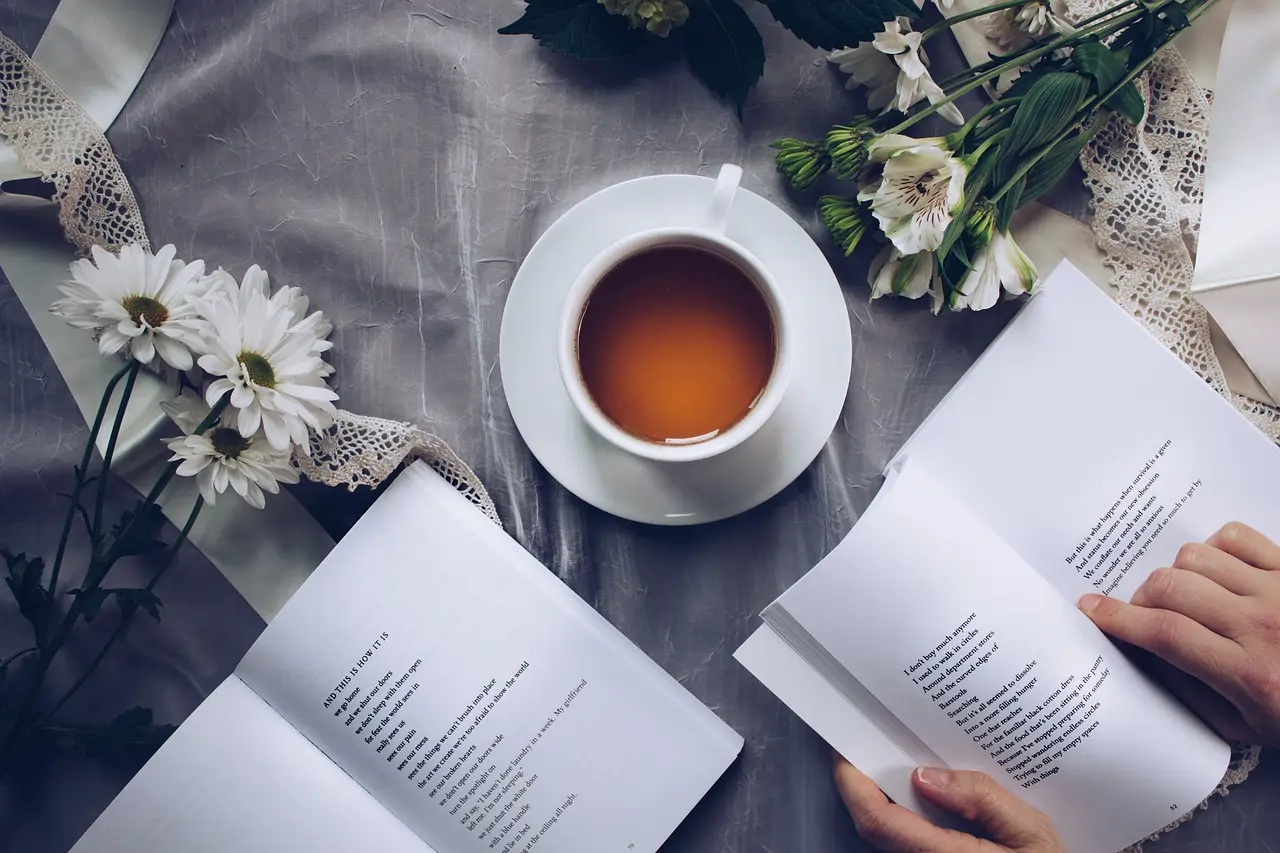 Ein Buch wird gelesen, daneben steht eine Tasse Tee auf einer Tischdecke mit Blumen und Spitze.