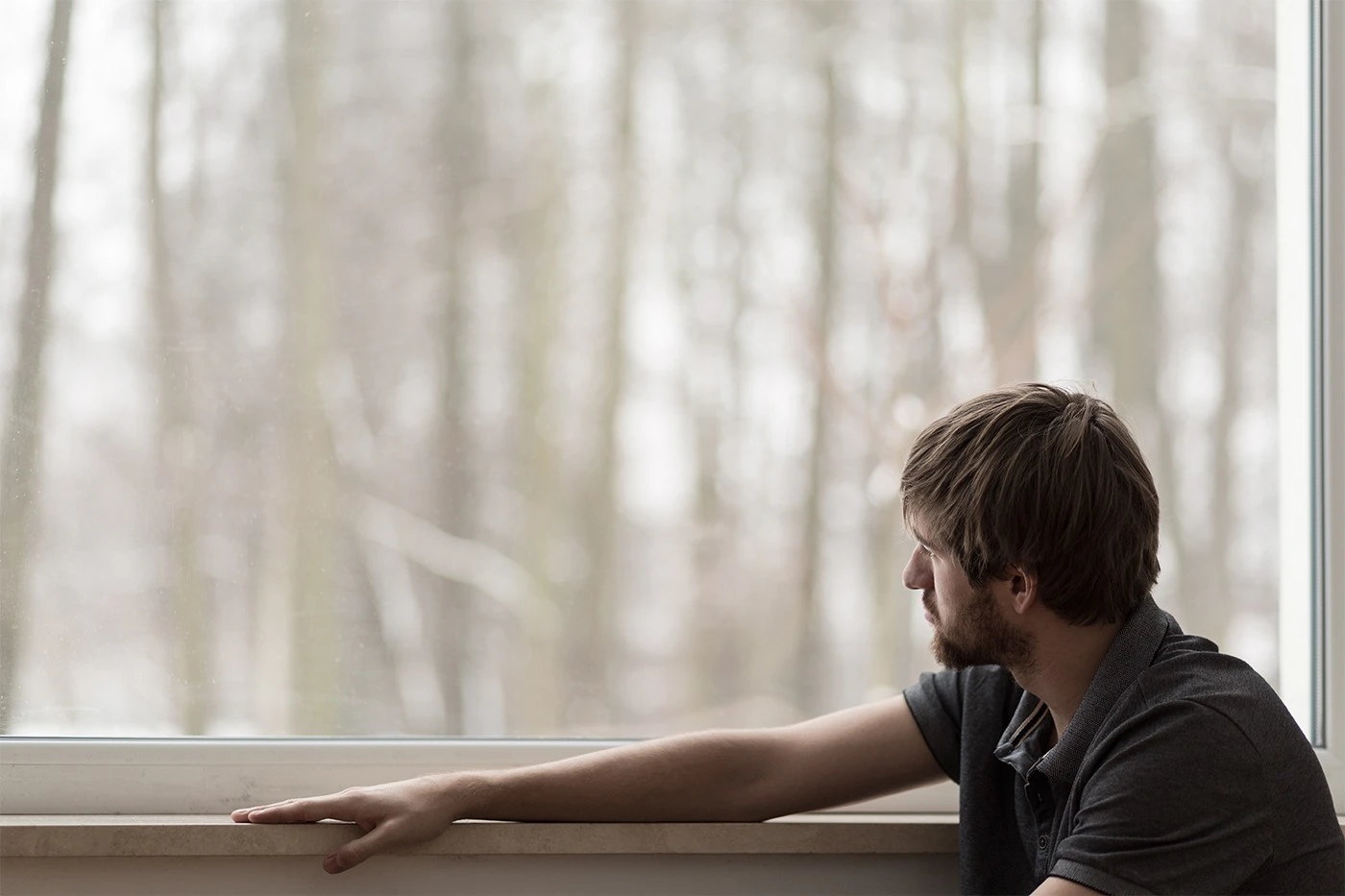 Ein nachdenklicher Mann sitzt an einem Fenster und blickt in einen nebligen, winterlichen Wald.